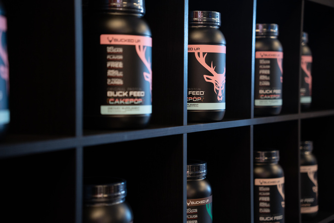 Several tubs of buck feed cake pop protein powder on a black shelf next to each other in individual cubbies.